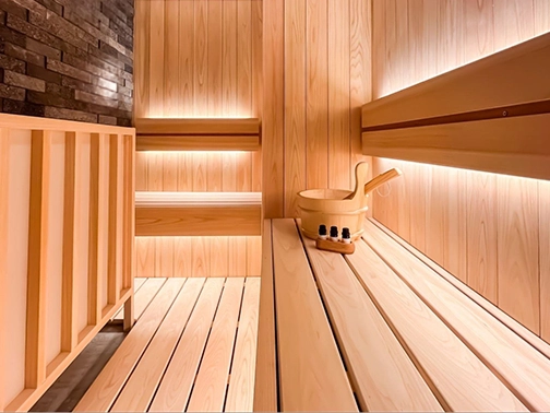 白木の壁と段差のあるベンチが美しいサウナ室、間接照明と黒いサウナストーブが配置された空間